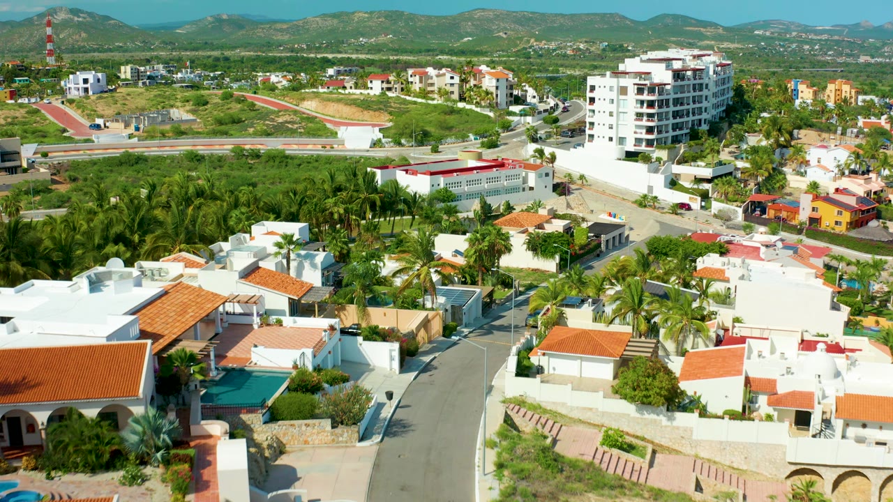 Opening aerial frame over San Jose del Cabo homes with white walls, red-tile roofs, palm trees, a curving road, distant hills, and no visible people.
