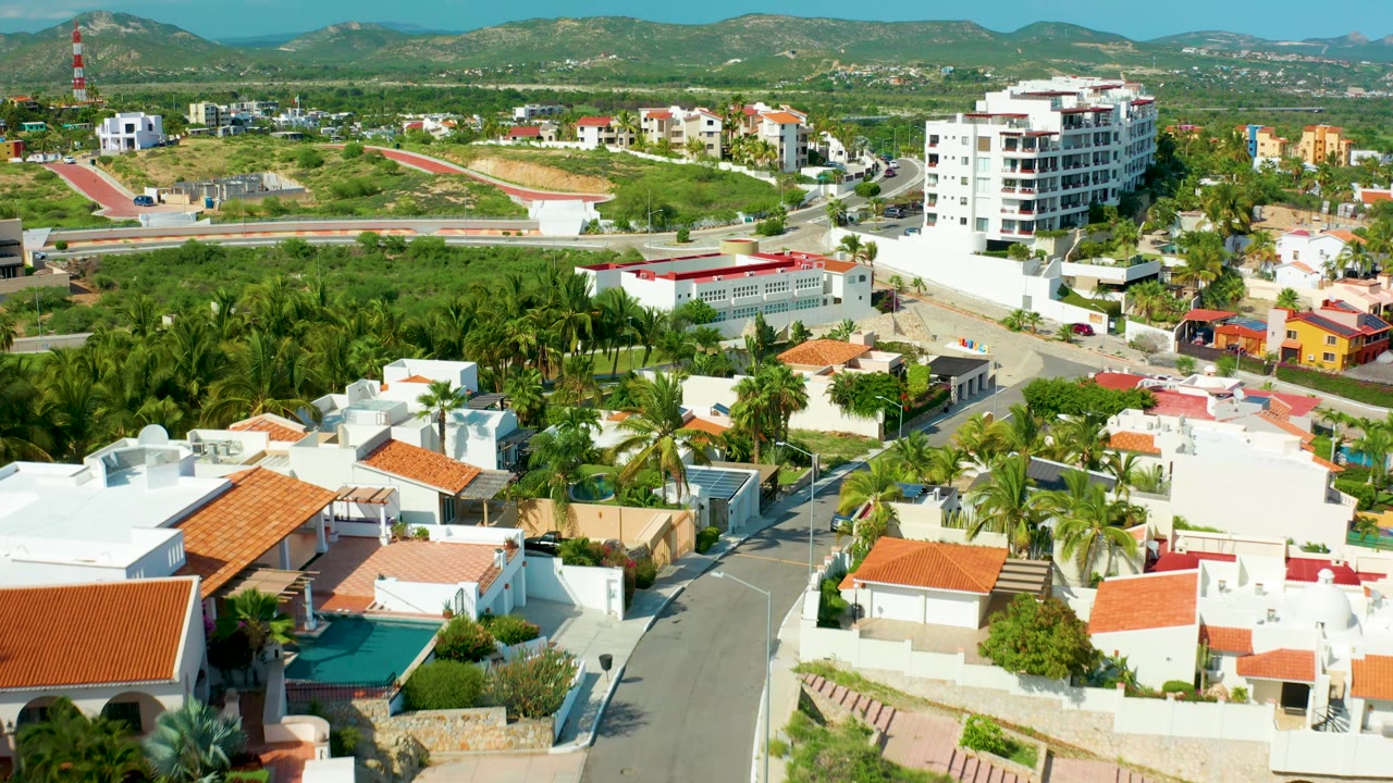 Aerial frame over San Jose del Cabo with white villas and red roofs in the foreground, a palm grove and road network in the midground, hills beyond, and no visible people.