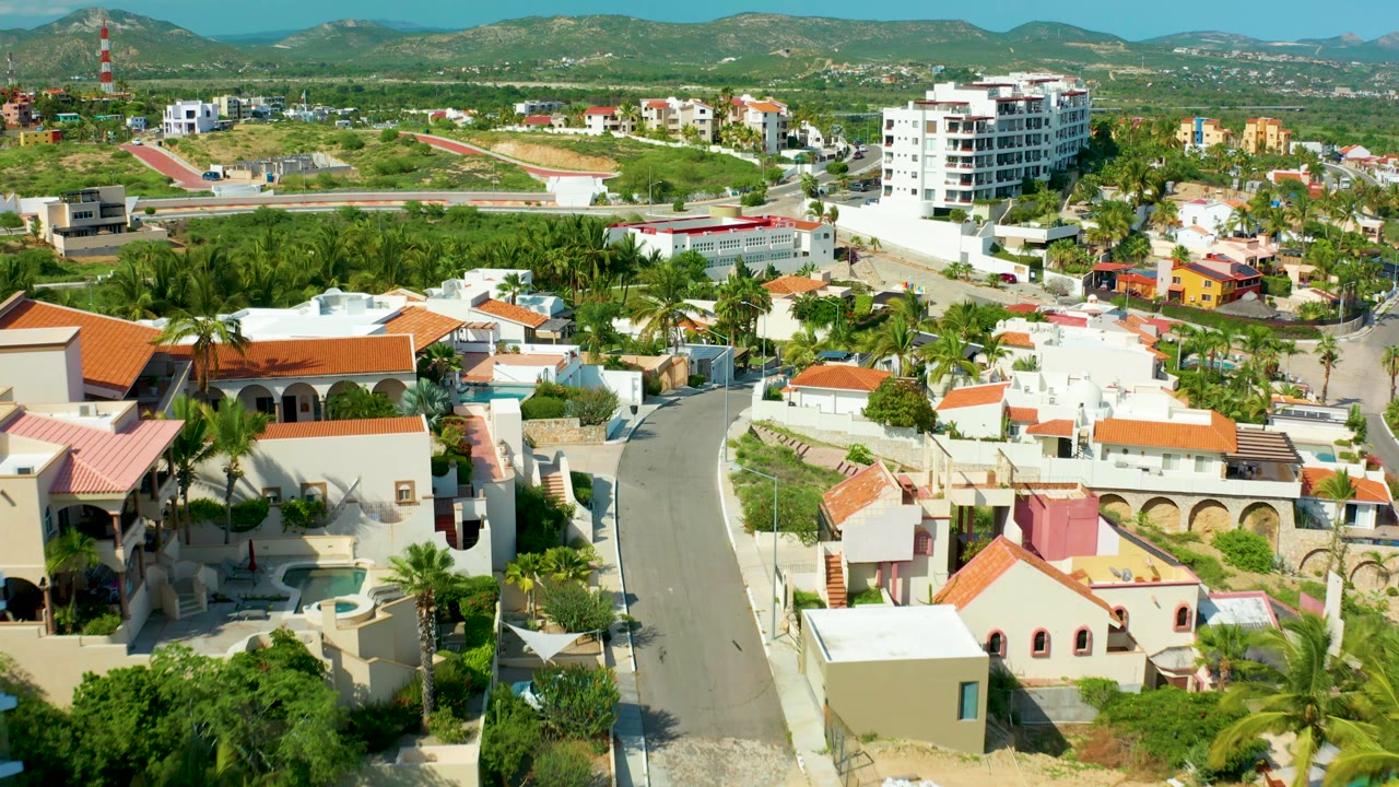 Opening frame of a San Jose del Cabo residential neighborhood with a curving road, white homes, orange tile roofs, palms, green hills, distant mountains, and no visible people.