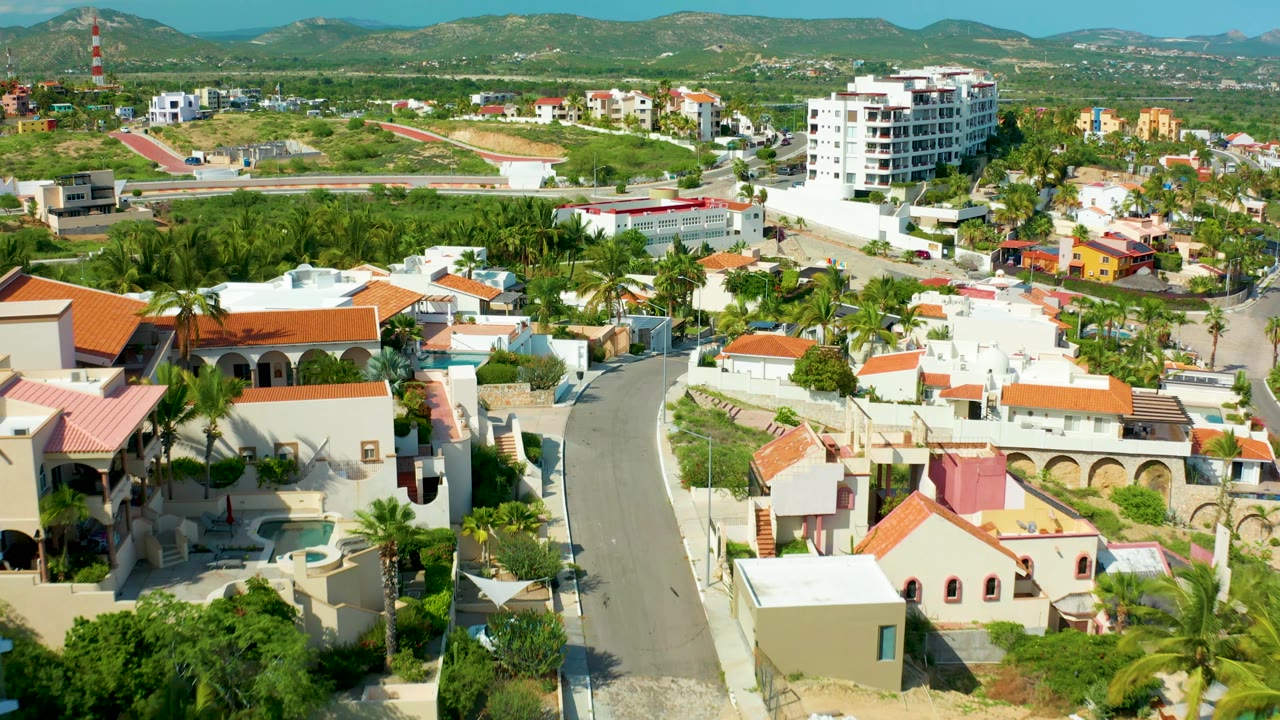 Early frame over San Jose del Cabo homes where the curving road runs between white villas, orange roofs, palms, a mid-rise building, and mountain foothills, with no visible people.