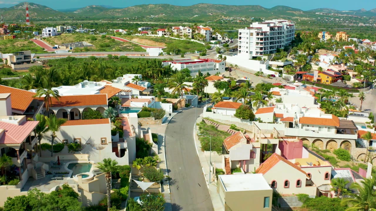 San Jose del Cabo aerial frame showing villas with orange and white roofs in the foreground, palms and a white apartment-style building in the midground, and mountains under clear sky, with no visible people.