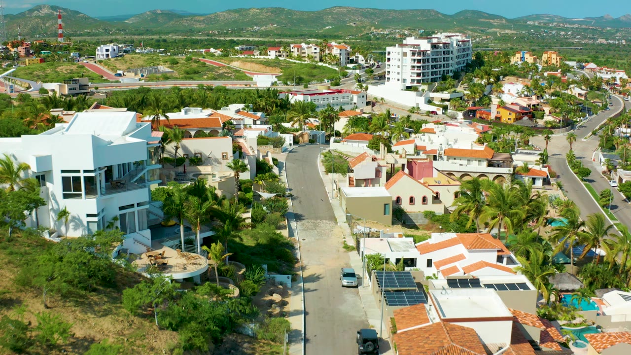 At 0:00, a drone view over San Jose Del Cabo shows white homes, red tile roofs, palm trees, a curving road, distant hills, and no visible people.