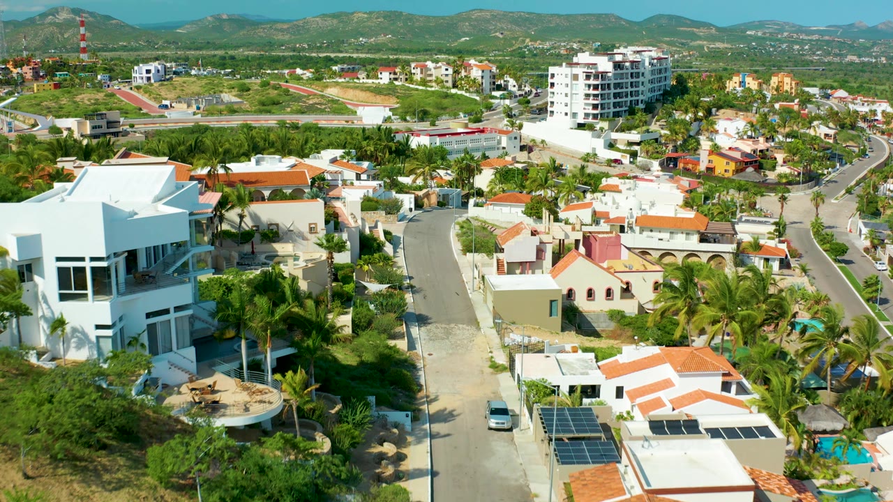 At 0:02, San Jose Del Cabo homes fill the frame with a large white house at left, palm trees below, a road through the center, hills behind, and no visible people.