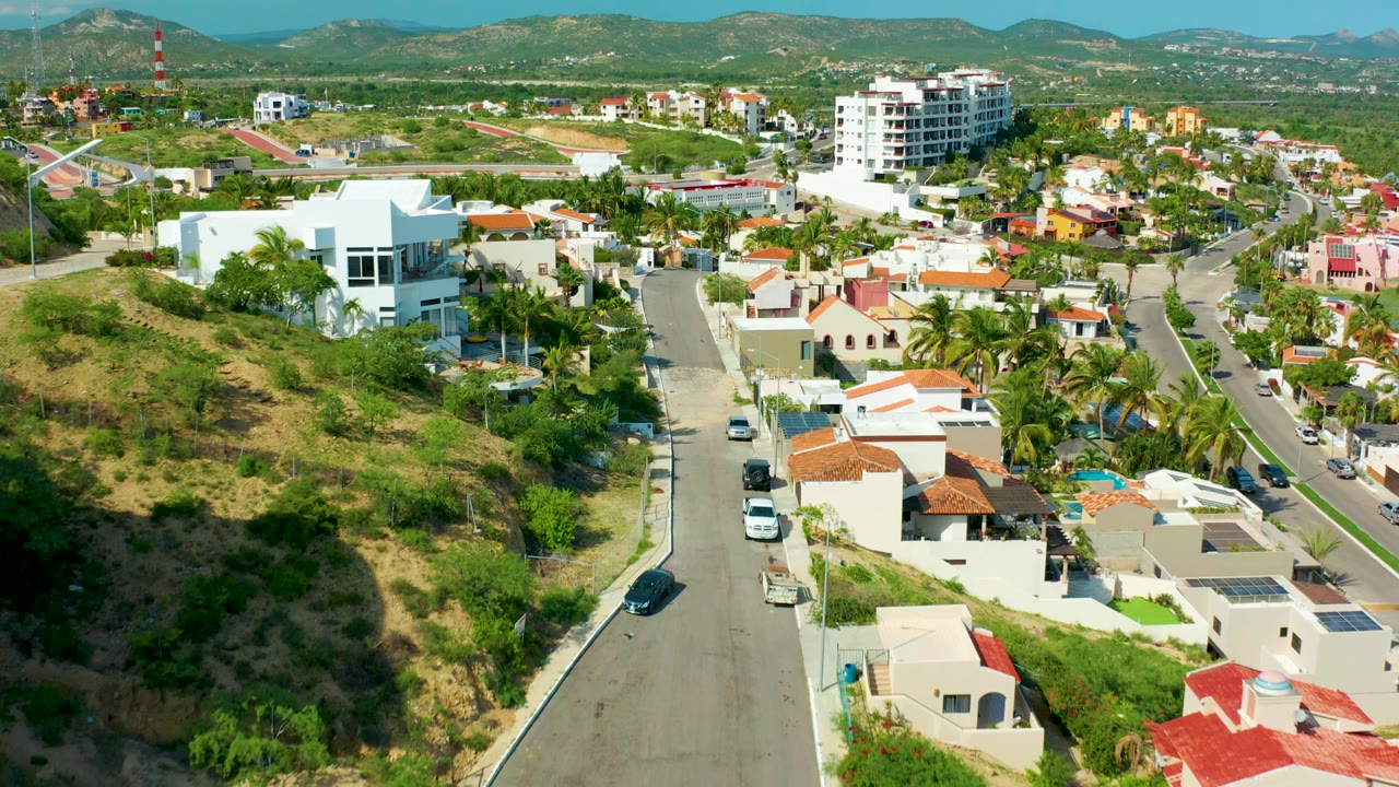 Aerial frame over San Jose Del Cabo hillside homes, with a residential road descending past white houses, red tile roofs, palm trees, distant mountains, and no visible people.