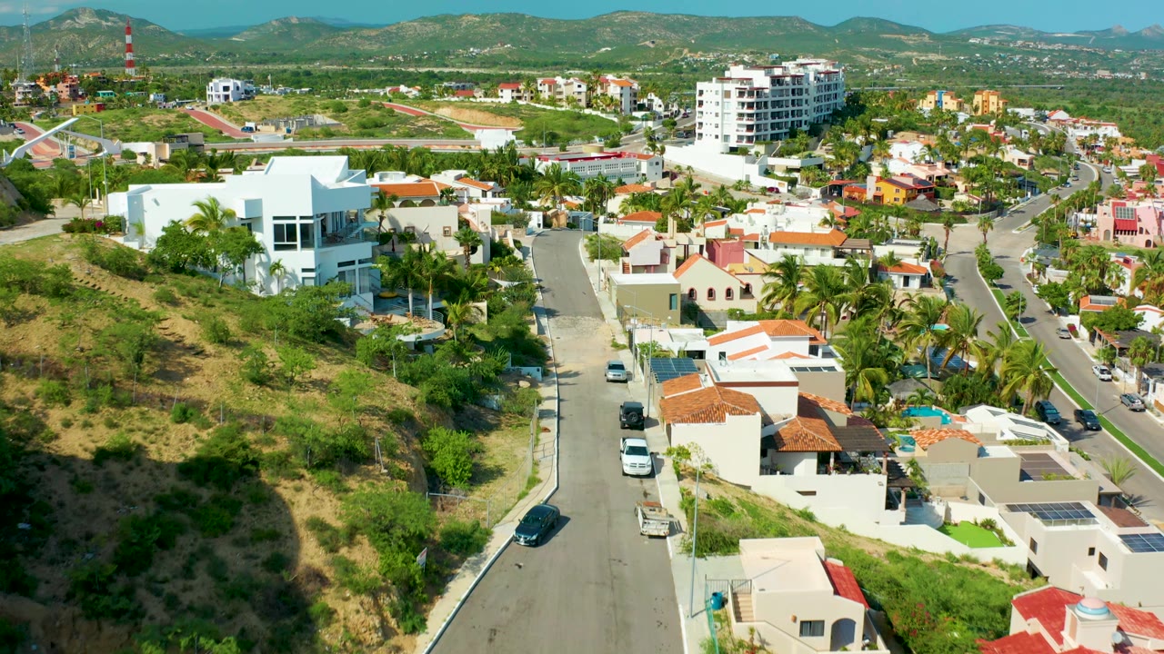 Aerial frame of San Jose Del Cabo homes clustered around a curving hillside road, with vehicles on the pavement, palms and shrubs around the houses, and no visible people.