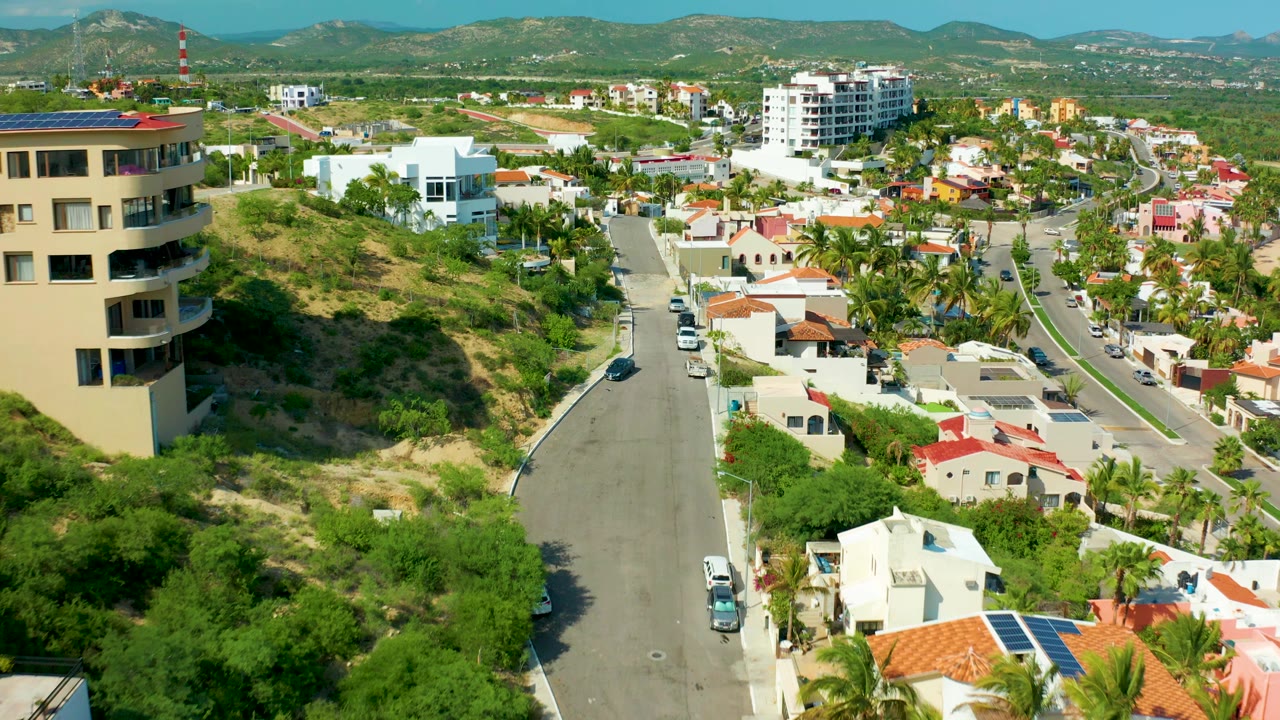 Opening aerial frame over San Jose del Cabo residential streets, with a tan rounded building on the left, red-roof homes, cars on the road, green hillside, distant mountains, and no visible people.