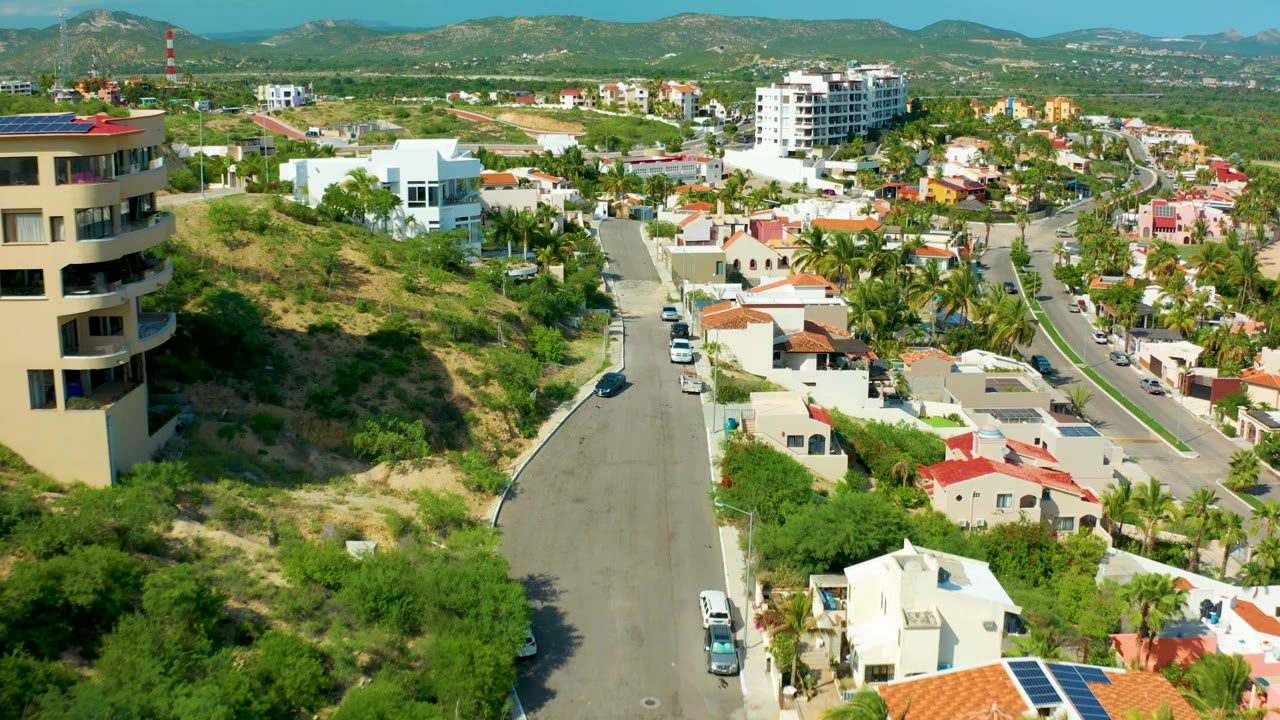 San Jose del Cabo aerial frame showing a curved residential road, houses with red and white roofs, parked and moving vehicles, a scrub hillside, a white multi-story building, distant mountains, and no visible people.