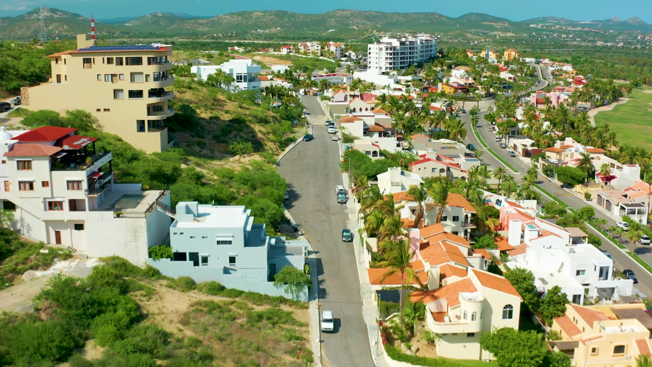 Opening aerial frame over San Jose del Cabo, Mexico, showing hillside houses, a paved road, green trees, distant mountains, and no visible people.