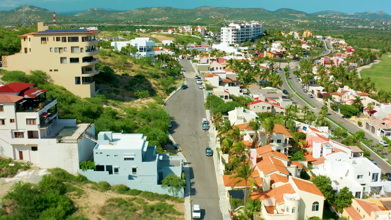 Aerial frame over San Jose del Cabo showing red-roofed homes, white and blue buildings, a curving neighborhood road, parked and moving cars, hills, and no visible people.