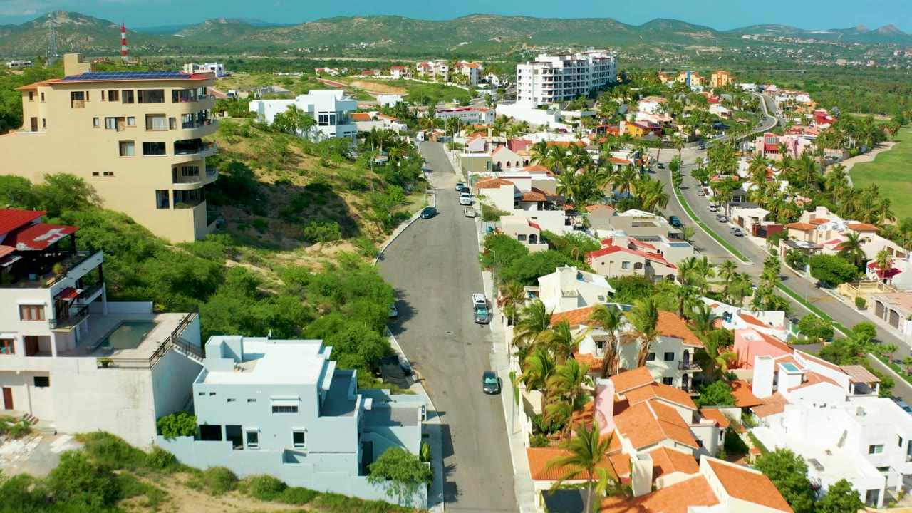 Aerial view of road between homes with beige building, white houses, trees, and hills.