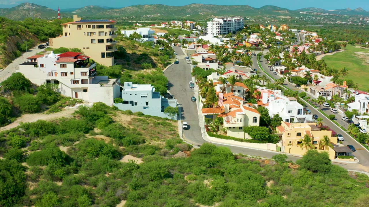 Aerial view one second in shows the beige hilltop building more centered above white homes and a winding residential road in San Jose Del Cabo, with no visible people.