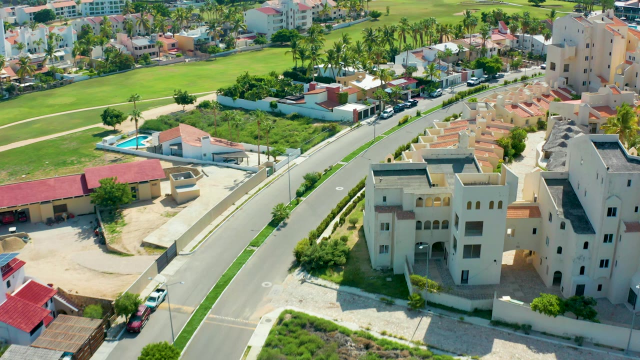 Aerial view in San Jose Del Cabo of a paved residential road curving past cream multi-story buildings, red roof homes, green fields, palm trees, parked cars, and no visible people.