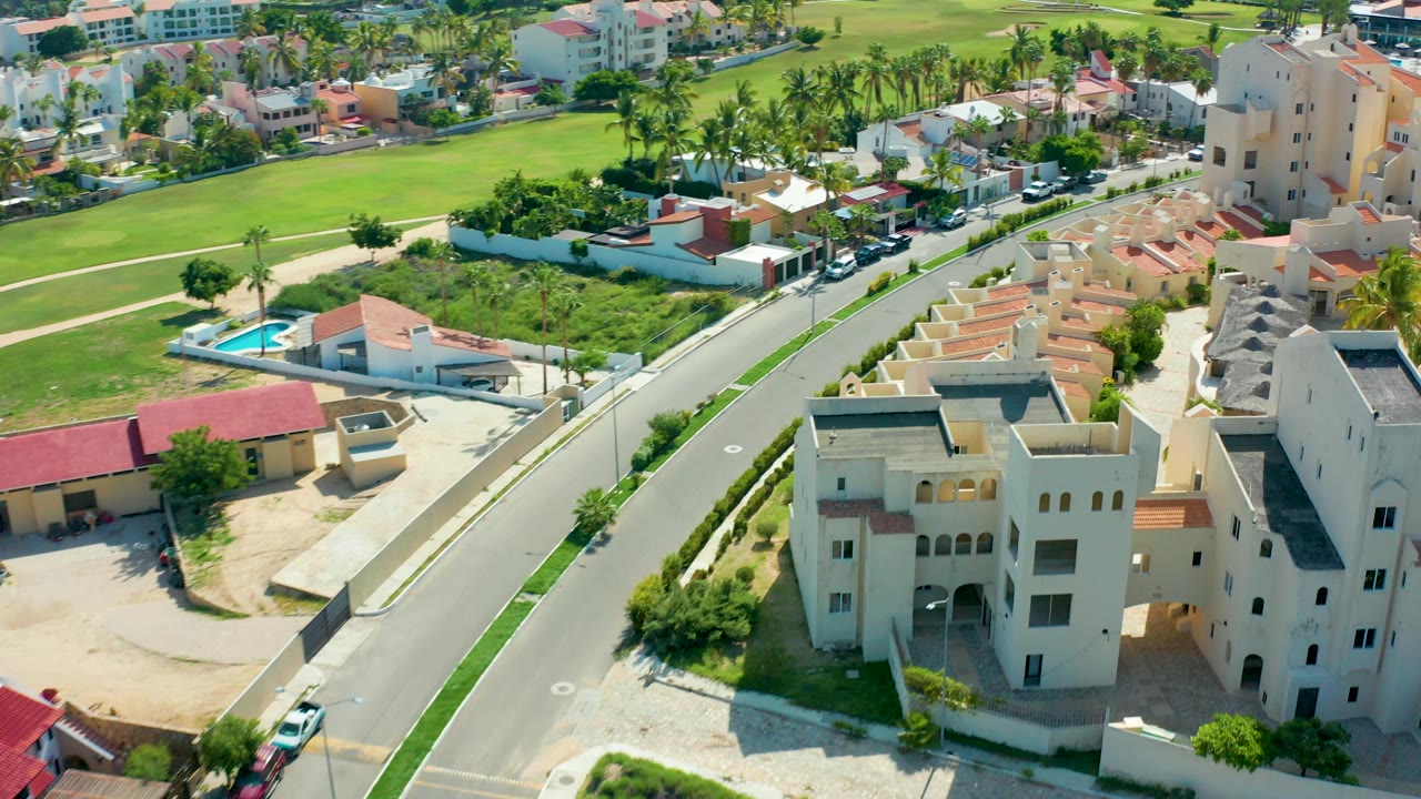 Aerial frame in San Jose Del Cabo shows the road sliding along cream residential buildings, with lawns and palm-lined homes in the background, parked vehicles, and no visible people.