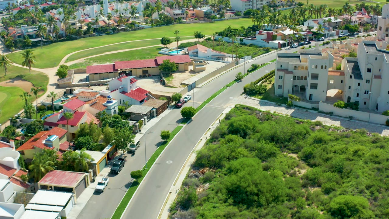 Aerial frame over San Jose Del Cabo showing a curved road, red-roofed homes, beige buildings, green open lots, palm trees, parked cars, and no visible people.