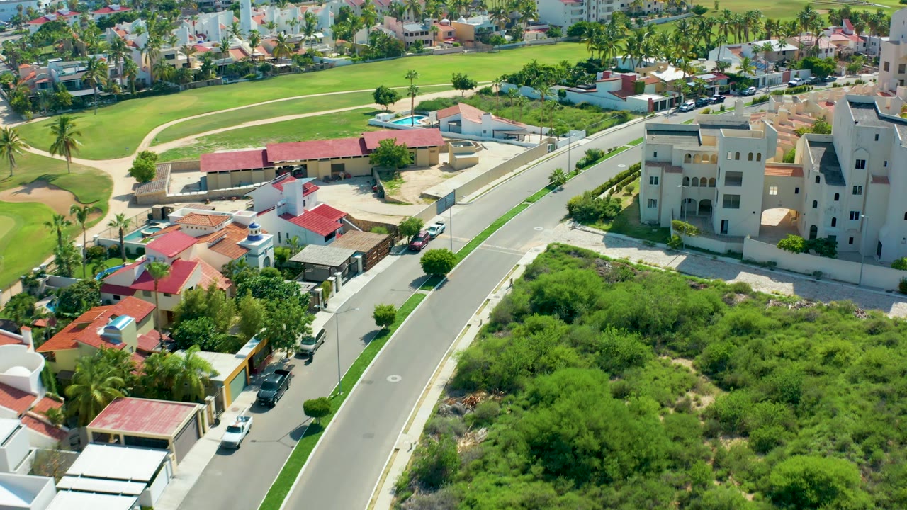 Drone view of houses, a road, parked cars, and green vegetation in a residential area.
