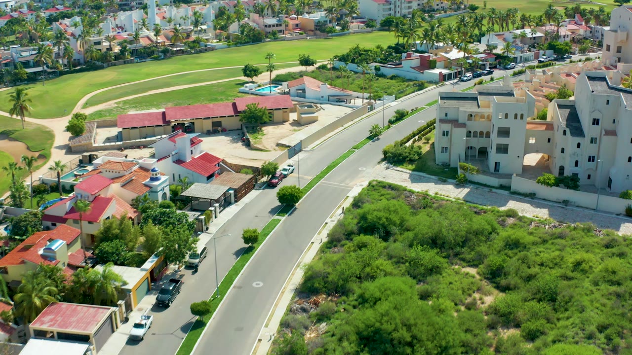 Aerial frame over San Jose Del Cabo showing more of the right-side beige buildings beside the curved street, with red-roofed houses, green parcels, parked cars, palm trees, and no visible people.