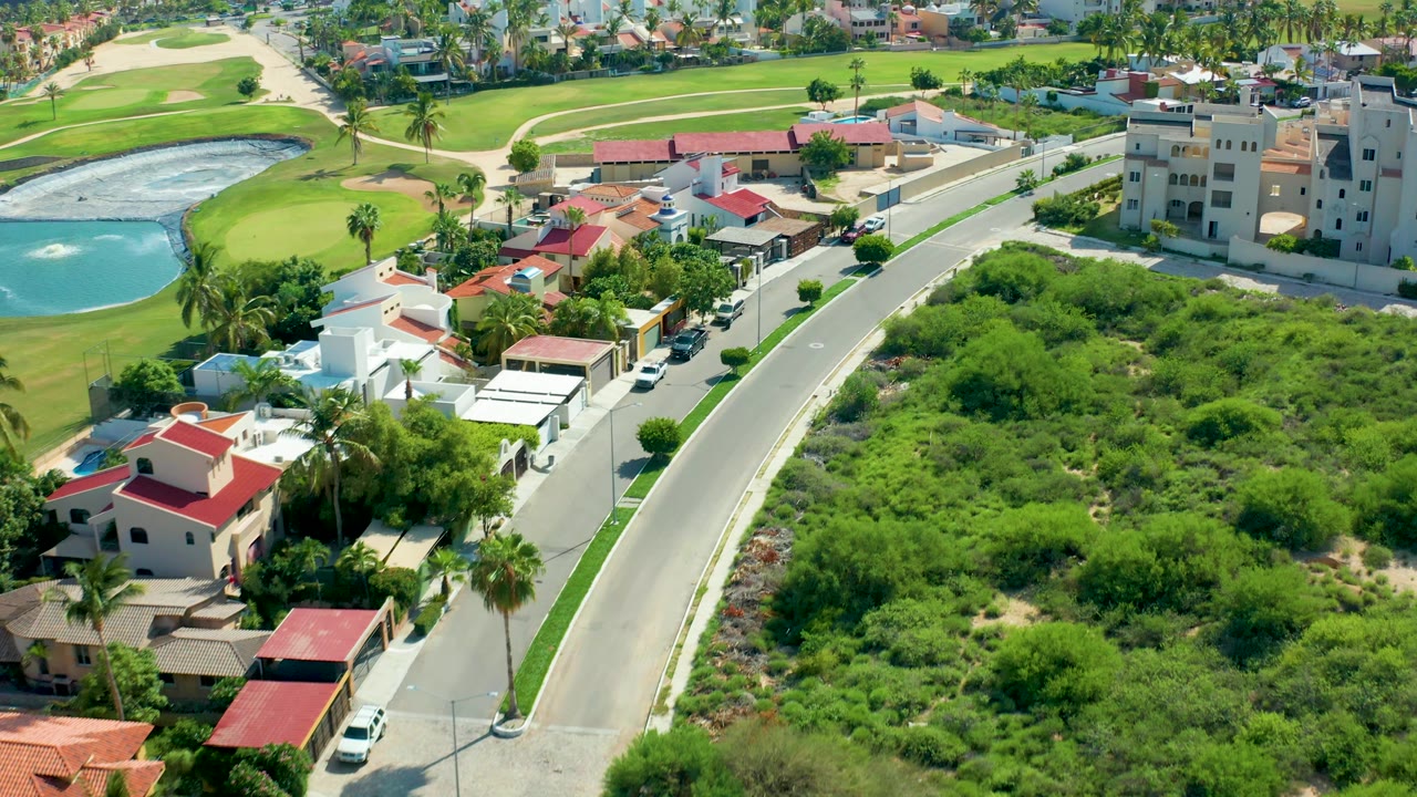 One second into the clip, the aerial view in San Jose Del Cabo centers the road between white and red-roof houses, parked vehicles, green brush, golf greens, and no visible people.