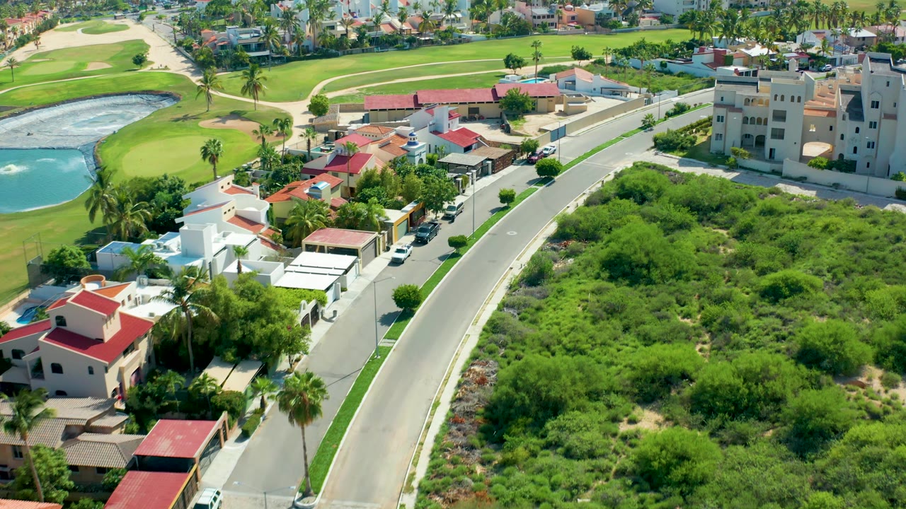 At 0:02, the San Jose Del Cabo aerial view keeps the curved road prominent, with dense vegetation filling the right side, houses and palms at left, golf course turf behind, and no visible people.