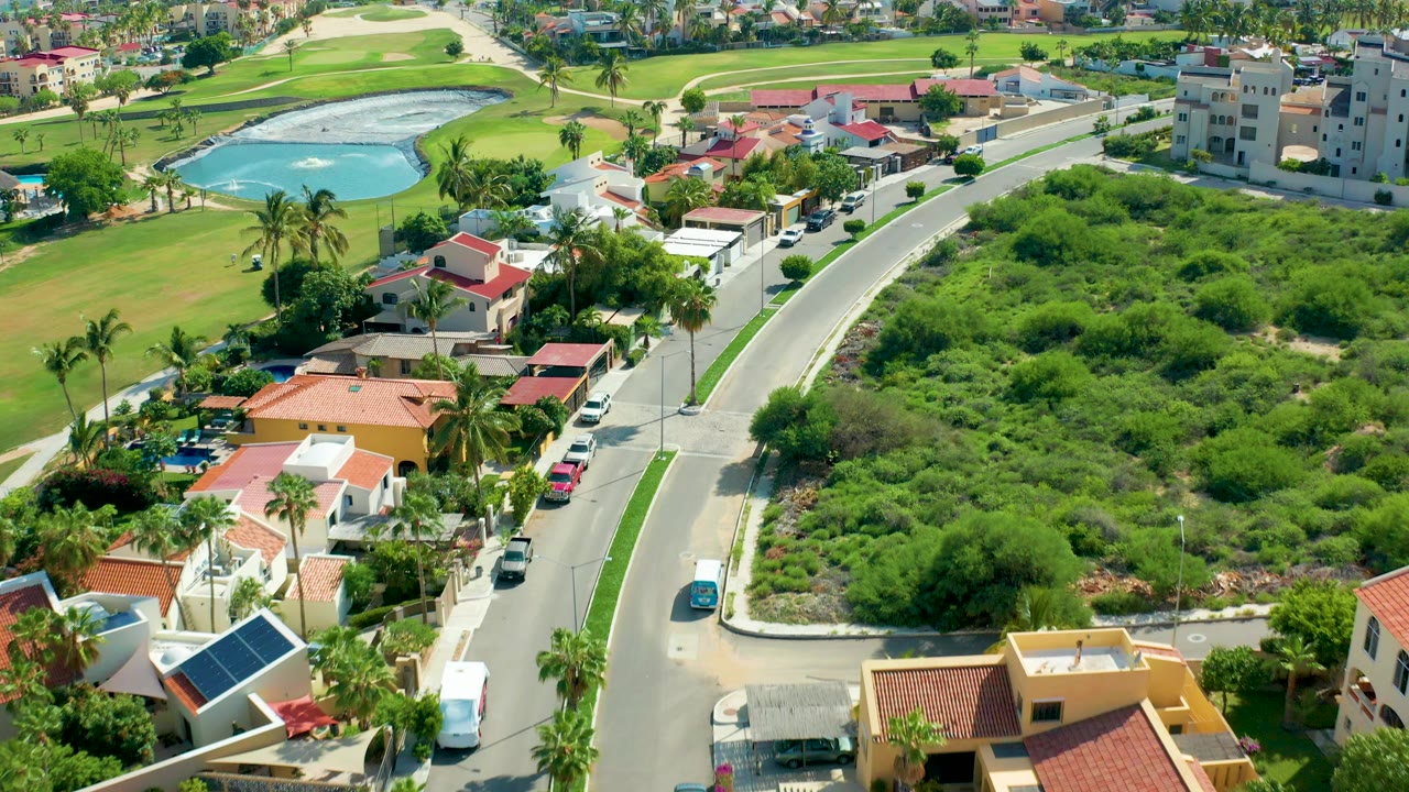 Opening aerial frame in San Jose Del Cabo showing a curved road beside red-roof homes, dense green vegetation, a golf course water hazard, and no visible people.