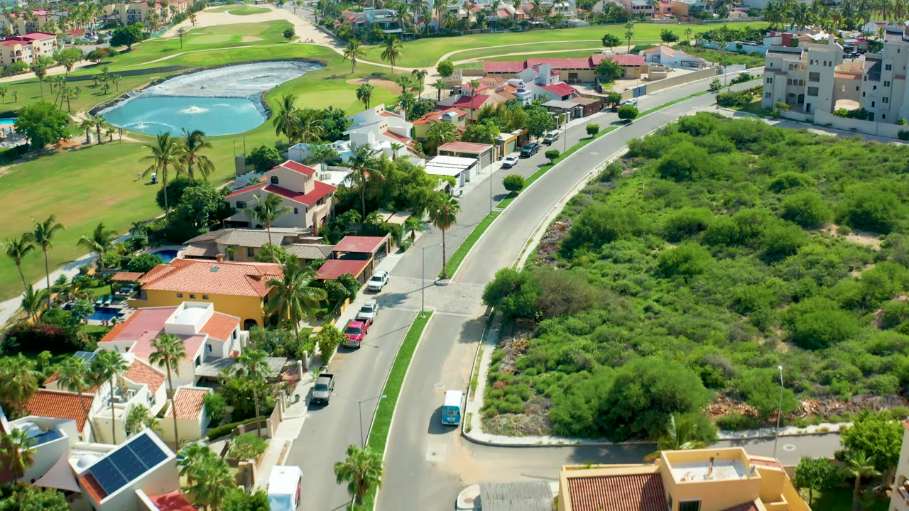 San Jose Del Cabo aerial view with a blue car on the road, villas and parked cars along the street, thick brush to the right, golf course water beyond, and no visible people.