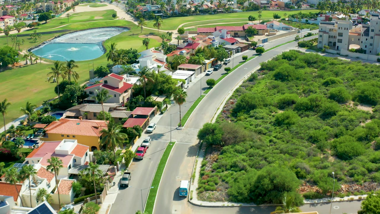 Aerial road bend next to dense vegetation, homes, palms, and golf course water.
