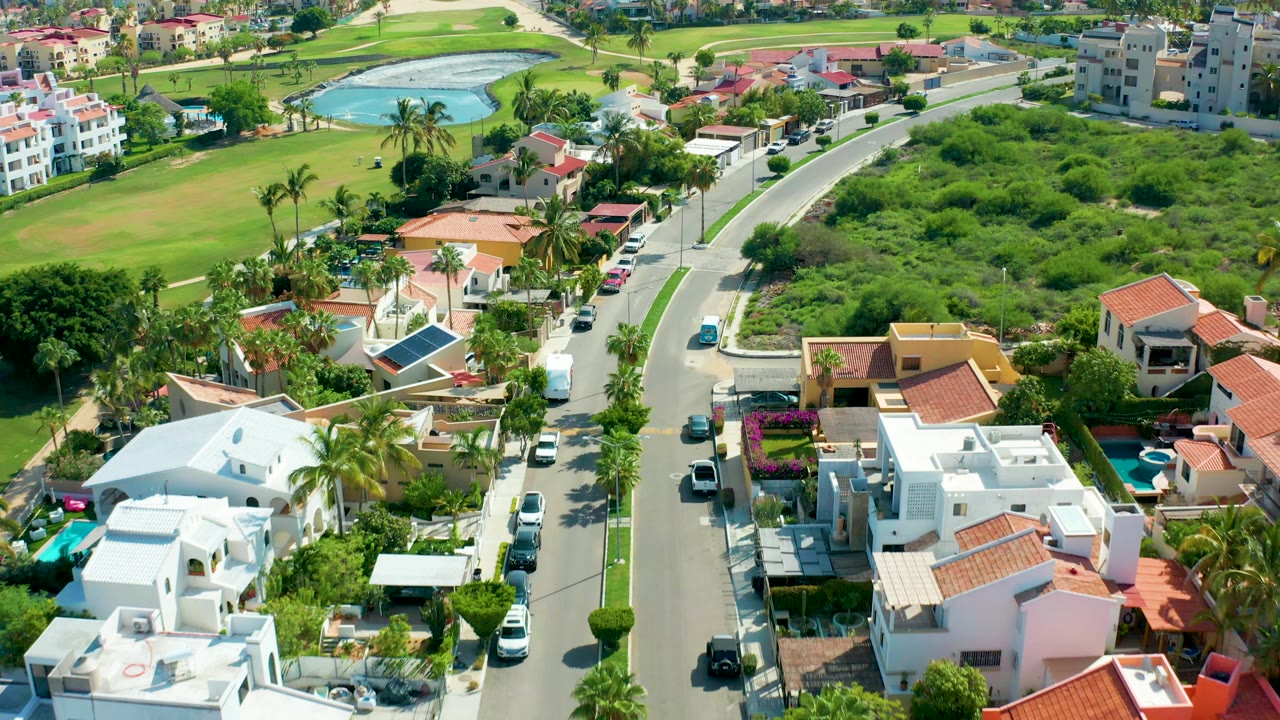Aerial frame from San Jose Del Cabo, Baja California Sur showing a curving residential street, white and terracotta-roof houses, parked cars, palms, green lots, a distant water feature, and no visible people.
