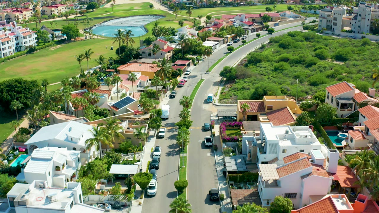Aerial frame from San Jose Del Cabo, Baja California Sur with the neighborhood road centered between white homes, parked vehicles, palm trees, dense green vegetation, and no visible people.