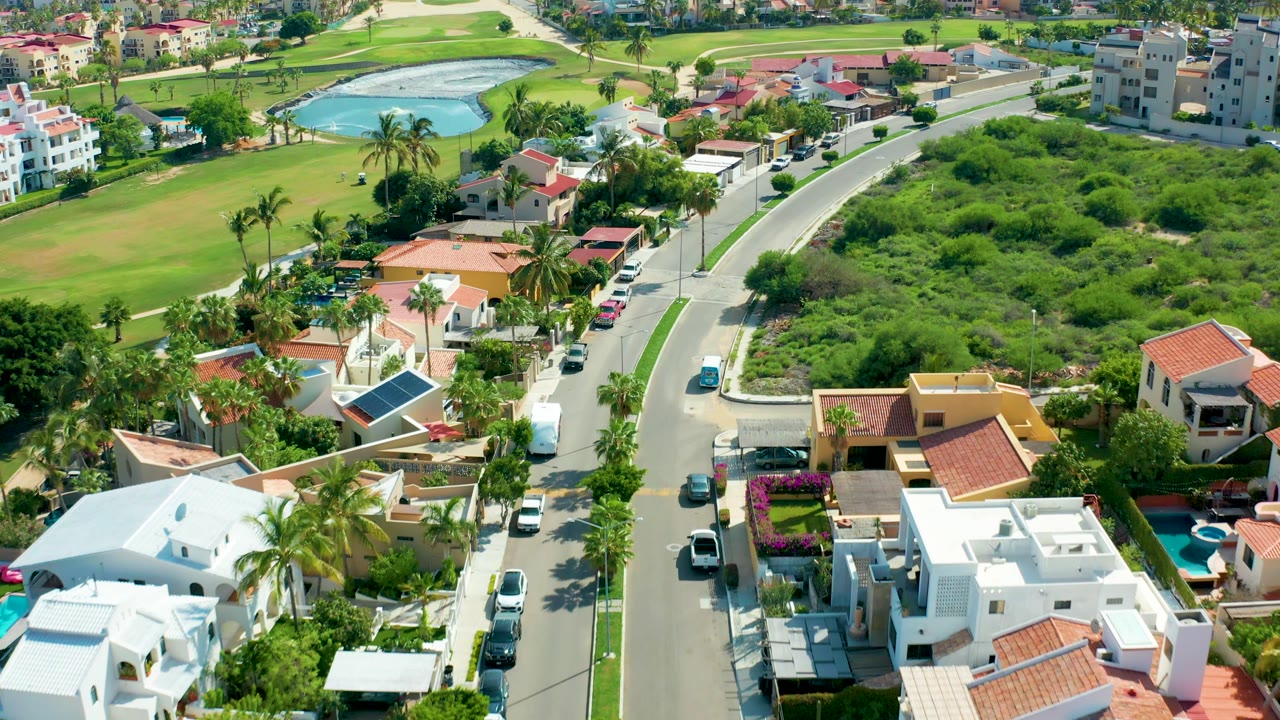 Drone-like view over houses and a street with parked cars, palms, a green field, and a water feature.