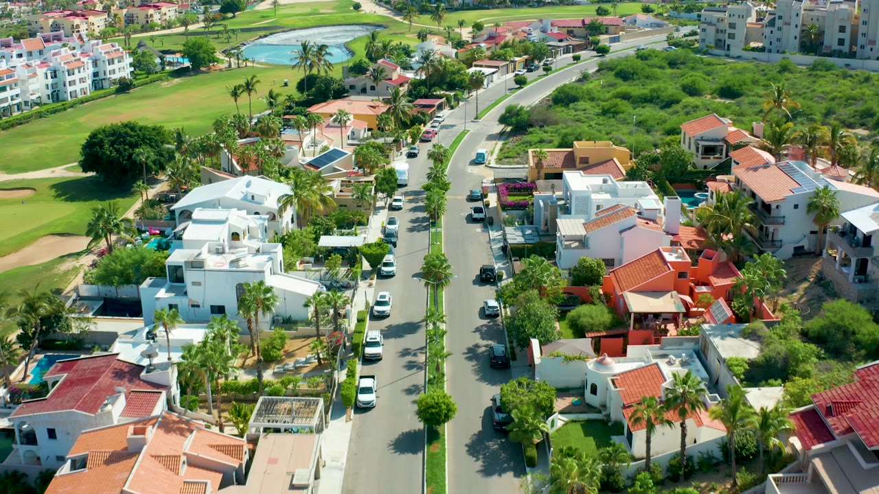 Drone view of a curved residential road between villas, palms, parked cars, and lawns.