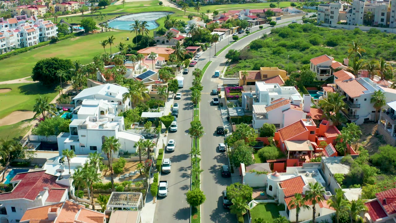 High aerial frame over San Jose Del Cabo with a red-roofed home prominent at lower right, white villas along the road, a blue pond in the distance, and no visible people.