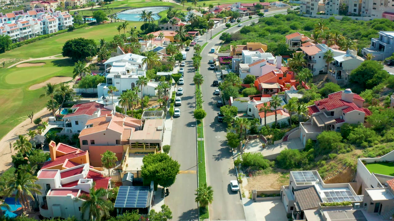 Aerial frame over San Jose Del Cabo showing a straight residential street between white and red-roofed homes, golf course grass on the left, parked cars, palms, and no visible people.