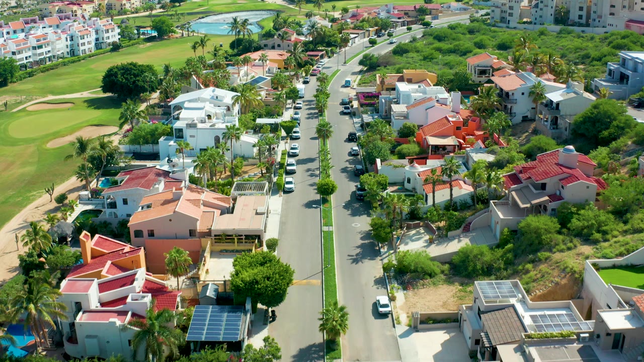 Aerial frame in San Jose Del Cabo with the camera view slightly farther along the street, showing white houses near the center, red and peach roofs, golf course turf at left, and no visible people.