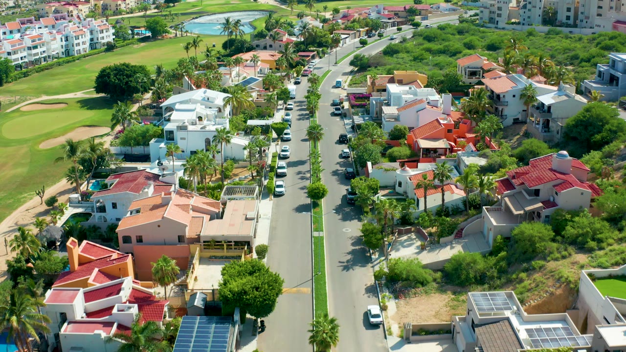 San Jose Del Cabo aerial frame showing red-roofed homes closer to the lower right, a line of parked cars along the street, golf course greenery on the left, apartment buildings in the distance, and no visible people.
