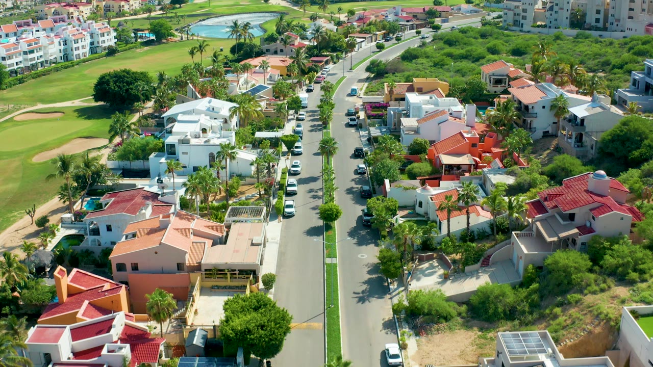 Drone-like view over a residential road with villas, golf course, and parked vehicles.