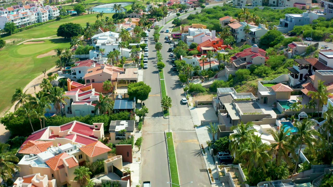 Drone-like overhead frame in San Jose Del Cabo showing the road running upward through homes, parked and moving cars, golf course fairway edges, palms, and no visible people.