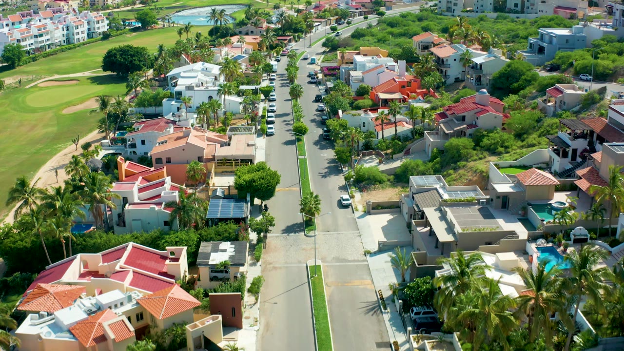 Aerial frame of San Jose Del Cabo residences with red and cream roofs, a straight road lined by palms, green golf course turf to the left, distant apartment buildings, and no visible people.