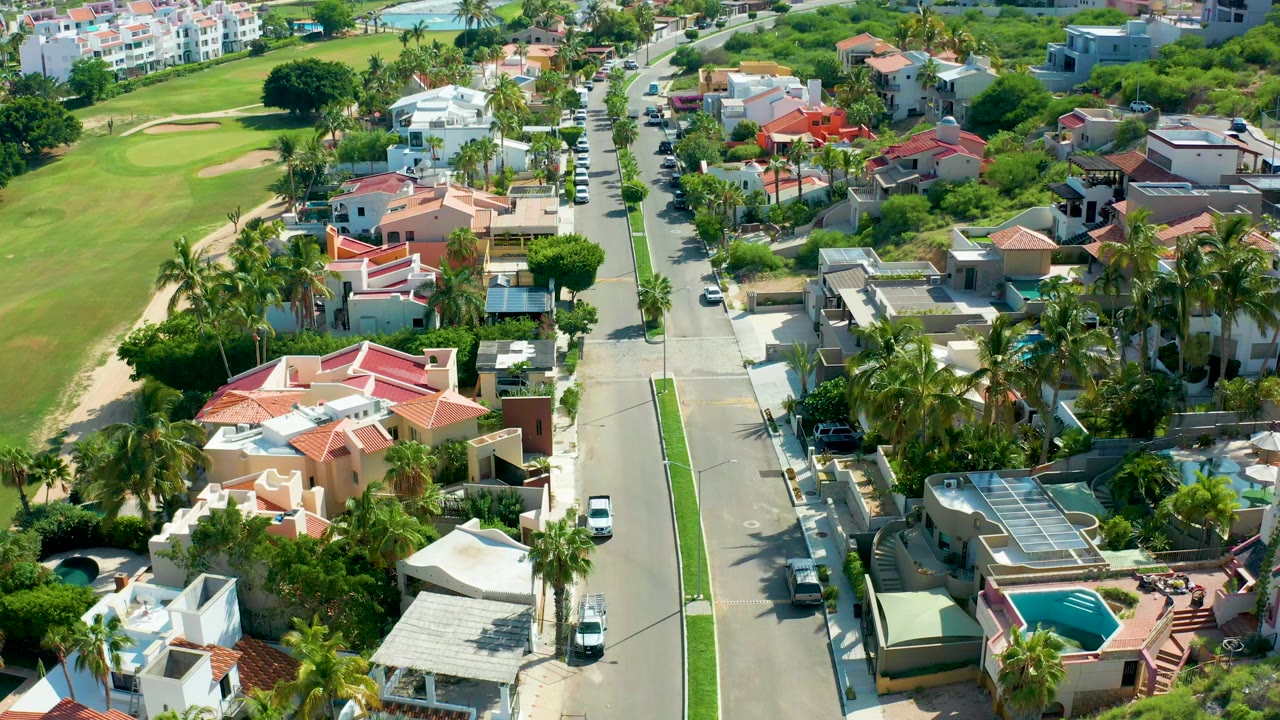 Overhead view in San Jose del Cabo, Mexico, showing a straight residential street with a green golf course on the left, private homes on both sides, parked vehicles, and no visible people.