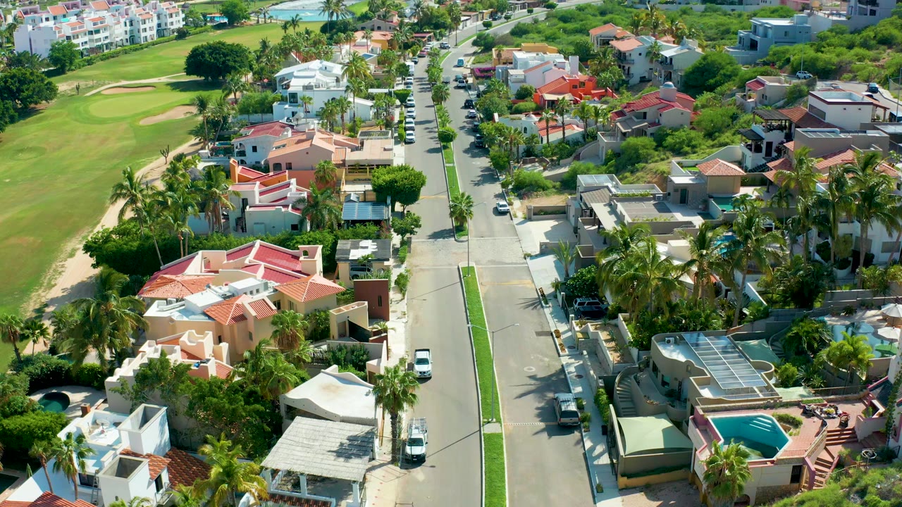 Elevated San Jose del Cabo street view with a white car lower on the road, a dark vehicle farther up the opposite lane, villas with red roofs, palms, and no visible people.