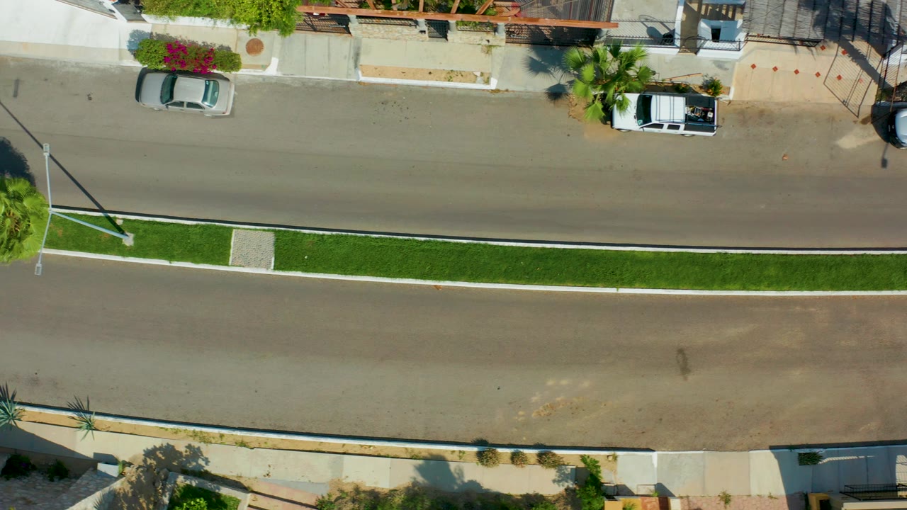 Top-down aerial frame in San Jose del Cabo shows a silver car near the left side of a quiet residential street, a grassy median below it, a parked white truck near the upper right curb, and no visible people.