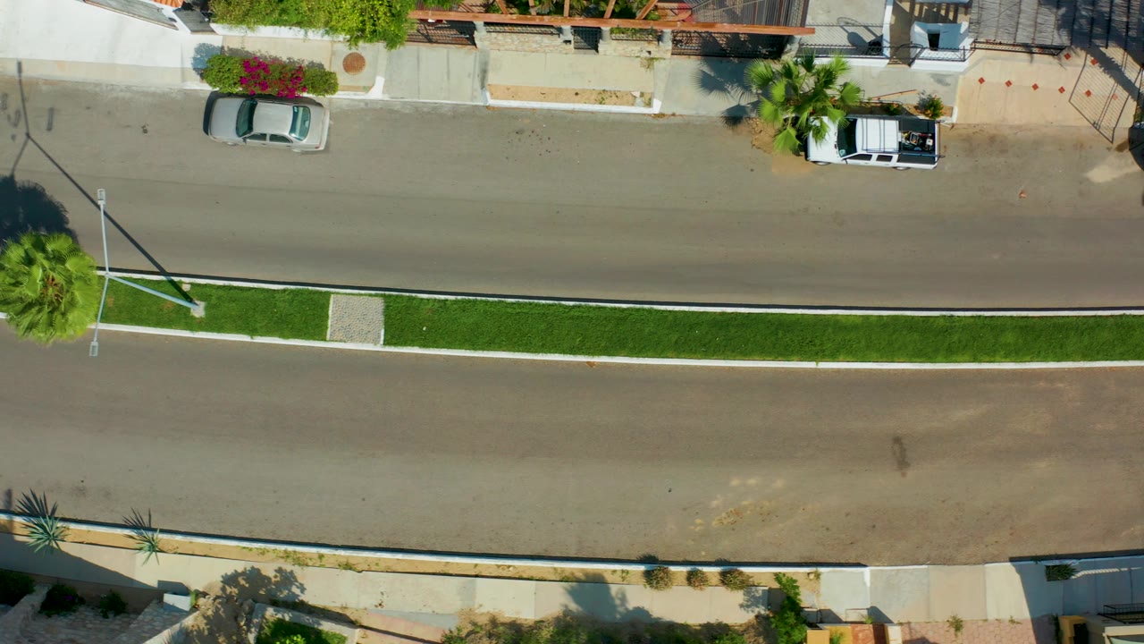 At 0:02, the top-down San Jose del Cabo frame places the silver car along the upper lane near curbside homes, with the parked white truck ahead, landscaped median below, and no visible people.