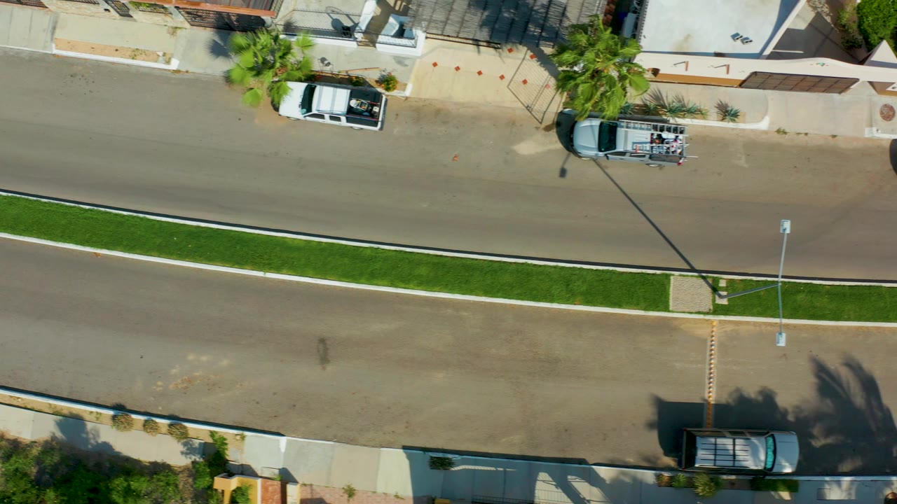 Straight-down view in San Jose Del Cabo showing the residential road shifted along the median, with a white vehicle by the curb, a dark vehicle at the lower sidewalk, palm shadows, and no visible people.