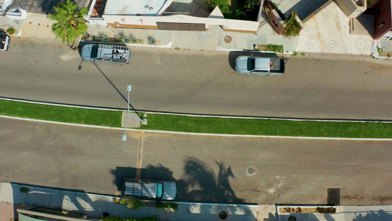 Early overhead frame in San Jose Del Cabo, Mexico, showing a white SUV near the upper curb, a parked blue car by the lower curb, the center grass median, and no visible people.
