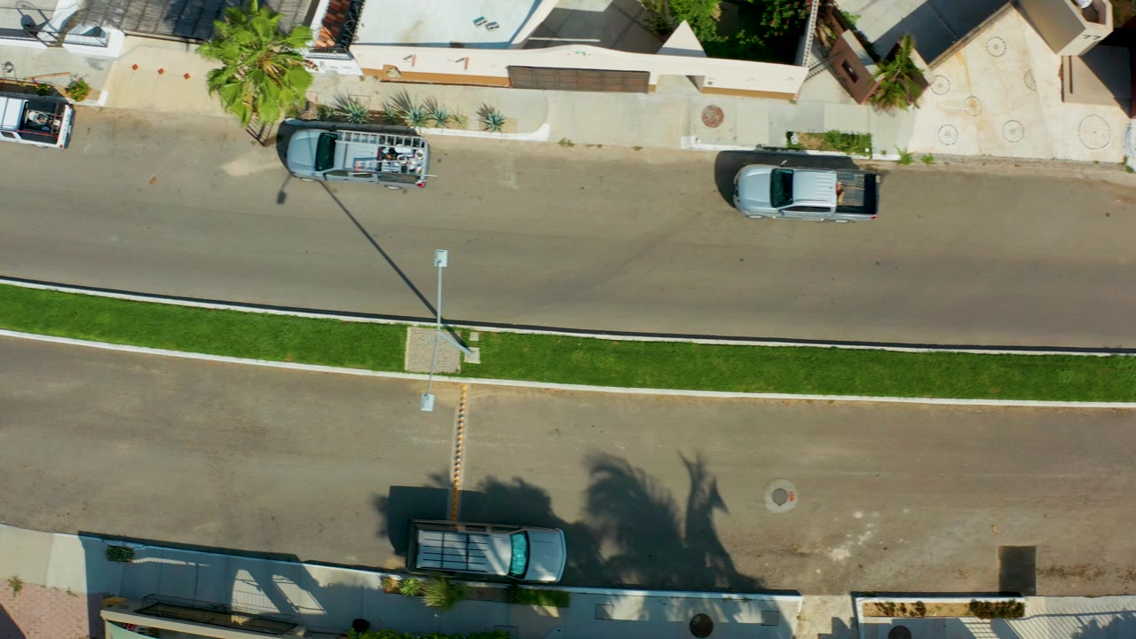 Overhead San Jose Del Cabo street view with the white SUV shifted farther right near the homes, the dark sedan on the road, the grass median below, and no visible people.