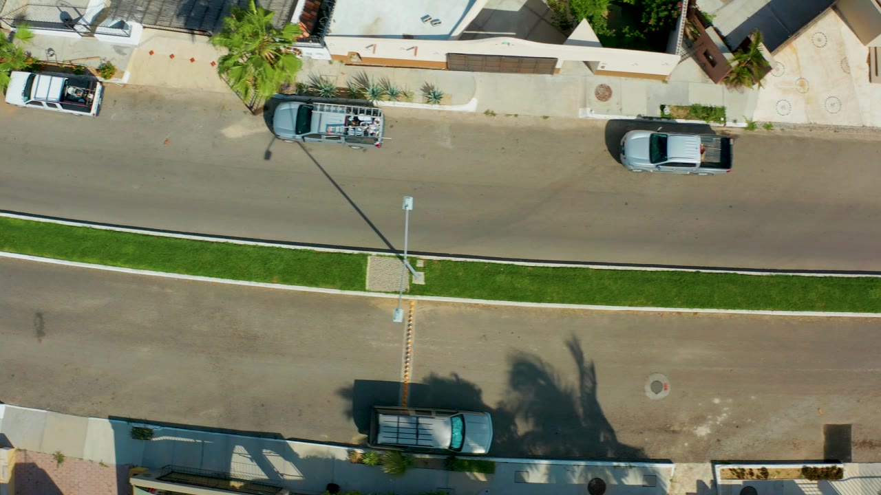 Residential street from above with a white SUV, dark sedan, parked car, and palm shadows.