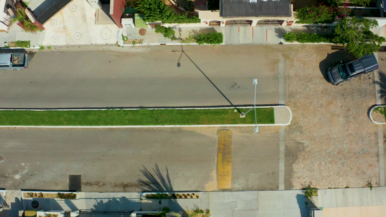 Overhead frame in San Jose del Cabo showing a residential street, grass median, pale homes, a light blue pickup at left, a dark vehicle near an upper driveway, and no visible people.