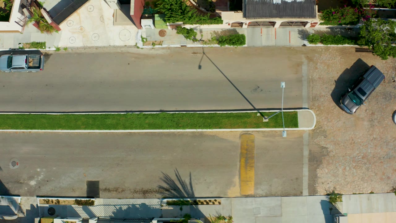 At 0:02, the overhead frame in San Jose del Cabo shows the dark vehicle angled near the right edge by gravel driveway space, the light blue pickup on the road, a grass median, and no visible people.