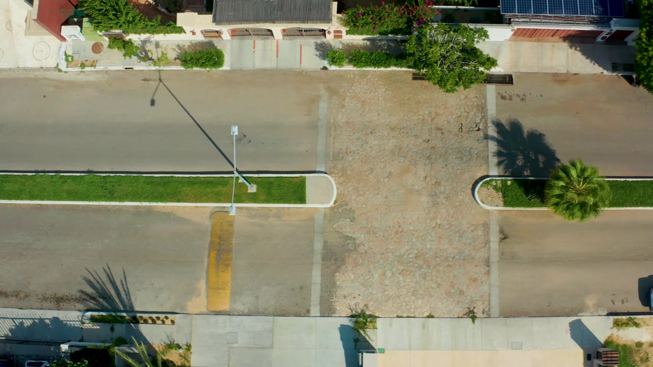 Overhead San Jose Del Cabo street view with the white vehicle at the right side of frame, a gravel driveway apron, planted median, sidewalks, and no visible people.