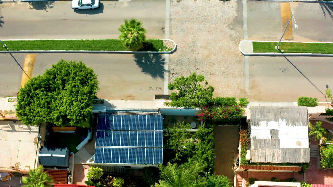Top-down San Jose Del Cabo view showing the white car near the top edge, a gravel lot, palm shadows, solar panels, residential roofs, and no visible people.