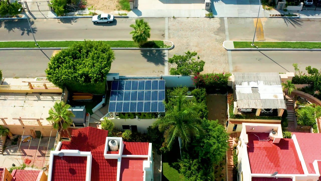 Overhead frame from San Jose Del Cabo showing red-roofed homes, a rooftop solar panel array, palm trees, a large shade tree, a road, and a white car near the upper street with no visible people.