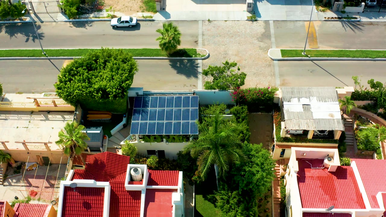 A high overhead San Jose Del Cabo frame shows the white car slightly farther along the road above red roofs, solar panels, palm trees, garden walls, and hard sun shadows with no visible people.